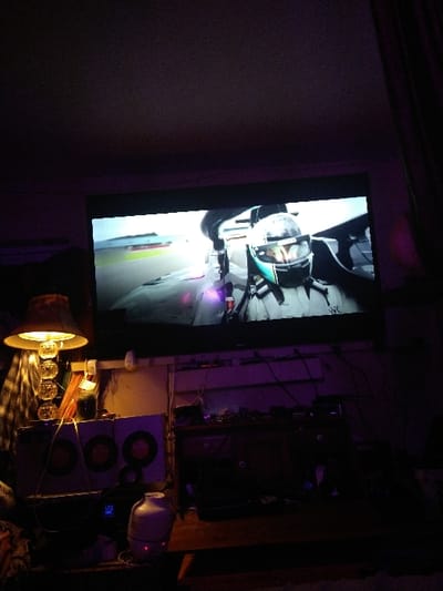 A dimly lit room features a large screen displaying a person in a helmet, possibly riding a vehicle. A complex network of wires and electronics is visible to the right of the screen, along with stacked speakers, a white lamp, and a horn-shaped light fixture illuminating several clear round objects. The overall scene suggests a makeshift entertainment or technology setup in a somewhat cluttered environment.