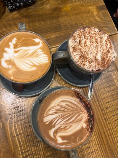 The image shows a tabletop view of three different types of beverages in mugs, likely coffee or hot chocolate, sitting on a wooden table. One mug is filled with a drink topped with a large amount of whipped cream and dusted with cocoa powder. The second mug has latte art featuring a feathered design. The third mug also has latte art. Each mug is on a saucer of the same color. A spoon rests on the saucer of the mug filled with whipped cream, and another spoon rests on the saucer of the mug with the feathered design latte art. A corner of a black and white patterned cloth is visible on the lower left. The photograph was likely taken indoors in Mole Valley, United Kingdom.