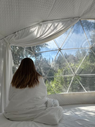 A woman with long, brown hair is sitting on a white bed, wrapped in a white duvet. She is facing away from the camera, looking out of a geodesic dome window. The window is made of clear panels framed by white metal structures. Outside the dome, there are trees and a blue sky with some clouds. The interior of the dome is white and quilted. Sunlight is streaming through the window, casting shadows on the bed. The scene is likely a glamping accommodation. The specific location in Nechkino, Russia, is not evident from the image.