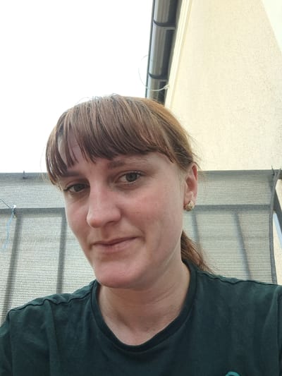This is a portrait of a woman with light brown hair and bangs, facing slightly to her left. She is wearing a dark green t-shirt. A subtle smile can be seen on her face, and she has a small, textured earring in her visible ear. The background shows a blurred outdoor scene with what appears to be a mesh-like structure or fence, and a light-colored surface. The lighting suggests it is daytime, possibly outdoors with bright, diffused light, indicating a clear or partly cloudy sky. The overall impression is a casual, candid selfie. No text is visible in the image. The location context provided is Mińsk Mazowiecki, Poland.
