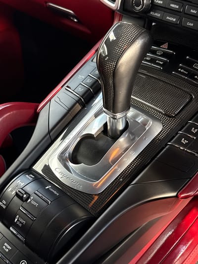 An eye-level shot captures the luxurious and functional center console of a car, identified as a Porsche Cayenne due to the prominent branding, within a setting of Blouberg, South Africa. The scene is well-lit, suggesting daytime. No people are visible.
The focal point is the automatic gear shifter, featuring a black grip with a textured, possibly carbon fiber, inlay. It is set within a sleek silver-toned gate displaying gear positions P, R, N, D, and M (manual mode). The word "Cayenne" is elegantly etched into the silver trim surrounding the shifter, which is itself embedded in a glossy carbon fiber weave trim that extends across the console.
To the left of the shifter, various infotainment and climate control buttons are visible, including "NAV," "CAR," "MEDIA," "PHONE," "TUNER," "SOURCE," "A/C MAX," and "SYNC," along with hazard lights and seat adjustment controls. Partially visible text includes "AUTO" and "REST."
On the right side, a large rotary dial and associated buttons manage driving modes, labeled "OFF ROAD," "ON ROAD," "comfort," "sport," and "sport plus." Additional controls for suspension settings, indicated by "H," "0," and "L" symbols, are also present, alongside a distinct "SPORT PLUS" button.
The interior showcases a striking contrast between the dark carbon fiber and black plastics and a rich, deep red leather or synthetic material on the console's side and possibly adjacent seating, contributing to the vehicle's sporty and elegant cabin aesthetic.