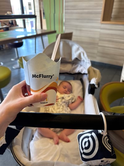 A hand is holding a McDonald's McFlurry container with a spoon inside. The container has the "McFlurry" logo and a small airplane graphic. Text on the side of the container reads "I'm lovin' it®". In the background, a baby is lying in a bassinet. The bassinet is black with white trim and a black and white patterned toy attached to the side. The scene appears to be indoors, possibly in a restaurant or cafe, given the presence of chairs and tables in the blurred background. The lighting suggests it is daytime. The overall tone is casual and relaxed.