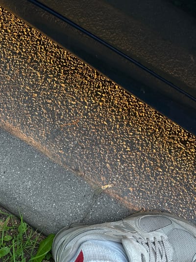 A close-up, low-angle shot captures a section of an asphalt surface next to a dark, horizontal object, possibly a car door, in Mārupe, Latvia. The asphalt is textured with small, dark grey pebbles. A diagonal line of golden sunlight illuminates the right side of the asphalt, creating a gradient of light and shadow. A thin, curved line of lighter grey pavement runs diagonally from the bottom left to the center, suggesting a change in surface material or a curb. In the bottom left corner, a patch of green grass and leaves is visible. The bottom right of the image features the upper portion of a grey athletic shoe with white laces, and a glimpse of a white sock with a red stripe. The scene appears to be outdoors, likely on a street or sidewalk, during a time of day when the sun is low, casting long shadows and warm light.
