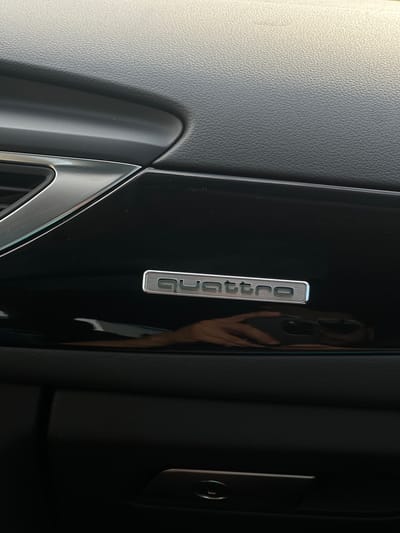 The image shows a close-up of the interior of a car, likely a dashboard. A metallic badge with the word "Quattro" is prominently displayed on a glossy black surface. Above this badge, the textured grey surface of the dashboard is visible, with a subtle reflective trim. Below the badge, a dark reflective surface shows a distorted reflection of a hand holding a phone, indicating the presence of a person taking the photograph. The lower part of the image reveals a dark compartment, possibly a glove box, with a metallic latch. The scene is indoors, suggesting the car is parked. There is no visible indication of the city of Sânnicoară, Romania.