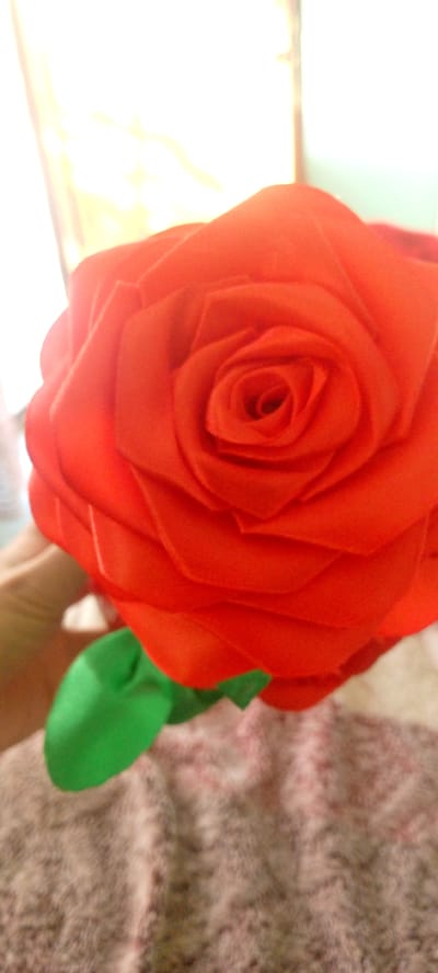This is a close-up, high-angle, indoor photograph of a bright red, artificial rose. The rose is centrally focused and dominates the frame, showing its layered petals in detail. The background is blurred and consists of a bright, diffused light source, suggesting a window or lamp, and a textured surface that appears to be fabric, possibly a blanket or upholstery. A hand, holding the stem of the rose, is visible in the lower left corner of the image. The stem is adorned with a green ribbon or leaf. The image does not contain any information or context that would place it within the city of Marhamat, Uzbekistan.