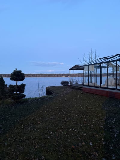 The image depicts a landscape in Baltezers, Latvia, featuring a glass greenhouse structure adjacent to a body of water. The greenhouse is positioned on a wooden deck. To the right of the greenhouse, the ground is covered in grass, leaves, and twigs. The body of water extends into the distance, with a visible shoreline opposite the greenhouse. In the foreground, there are several small trees or bushes. The sky is a clear blue.