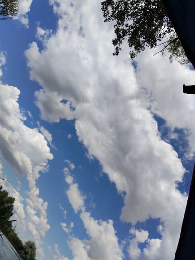 The image captures a sky with scattered cumulus clouds over a body of water. Trees and foliage are visible along the bank of the water. A dark blue and red edge of what appears to be a tent or awning frames the scene from the right. The lighting suggests it is daytime. The orientation of the image is tilted. The location is Bilyaivka, Ukraine, near the Dniester River.