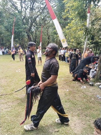In an open grassy field in Puro, Indonesia, a cultural performance is unfolding. A man with a darkened face, wearing black clothing with a yellow sash, holds a horsehair whip. Another man in a similar black outfit stands nearby. A crowd is gathered in the background, and red and white flags are placed around the field. Several other performers, also dressed in black, engage in some action on the ground. Tree foliage creates a backdrop to the scene.