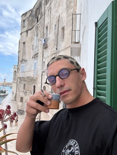 The image captures a close-up of a person on a balcony, observing a historic coastal town below. The subject is a young individual with short, light-colored hair, possibly bleached or patterned, and is wearing round, dark sunglasses. They are dressed in a black t-shirt featuring a partially visible white circular logo on the right side of the chest; text like "OLD LTD" and "201" are discernible within the logo, alongside a stylized creature. The person holds a lit cigarette in their right hand and a small, two-toned cup or container (with a terracotta base and grey top) in their left. Their facial expression is neutral.

In the foreground, green louvered shutters and a white balcony ledge are visible at the top, along with red succulent-like plants and green spiky plants in pots on the right. The background reveals a picturesque townscape, consistent with Monopoli, Italy, under a bright, partly cloudy sky. Weathered, light-colored stone buildings with numerous windows and balconies line a waterfront. Some windows have blue louvered shutters, and laundry is seen hanging to dry on one building. Below, a canal or harbor features at least one boat and a small lighthouse. Two small figures are visible relaxing on a lower level near the water.

The scene depicts a sunny, clear day, likely midday or early afternoon, with the person enjoying a moment of relaxation and observation from their elevated position.