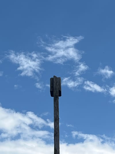 A close-up, low-angle shot shows a weathered wooden post reaching upwards into a bright blue sky filled with wispy white clouds. The top of the post is capped with four vertical wooden planks, forming a square, open-ended structure. The post is centrally located in the frame, with the sky dominating the upper portion and the bottom edge of the post visible. The overall lighting suggests daylight. This scene is unlikely to be in Pamyati 13 Bortsov, Russia, as the image lacks any contextual urban or geographical features.