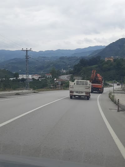 The image depicts a grey, overcast day with a mountainous, vegetated landscape. A paved road with a white lane marker curves through the scene. In the foreground, a white pickup truck with a flatbed is driving on the road. Behind it, a large orange truck with a crane attachment is also in motion. In the background, several buildings are visible nestled into the hillsides, along with a tall electrical pylon. The location is identified as being in Ayvacık, Turkey.