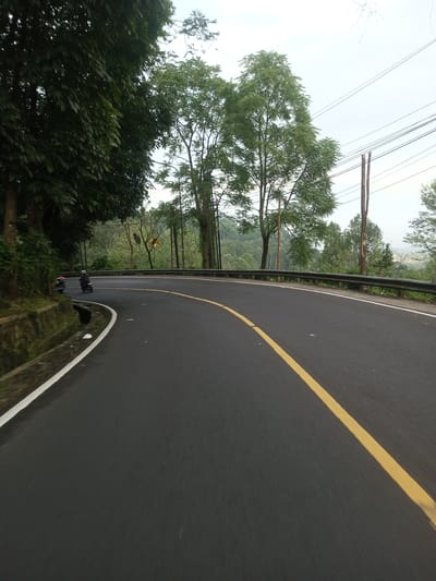 The image captures a curved asphalt road in Taunggyi, Myanmar. A solid yellow line marks the center of the road. On the left, there's a mossy stone wall alongside dense foliage. The right side features a guardrail with trees in the background and power lines overhead. Two motorcycles are faintly visible on the left side of the road. The sky is overcast. The road appears to be winding through a mountainous or hilly area.