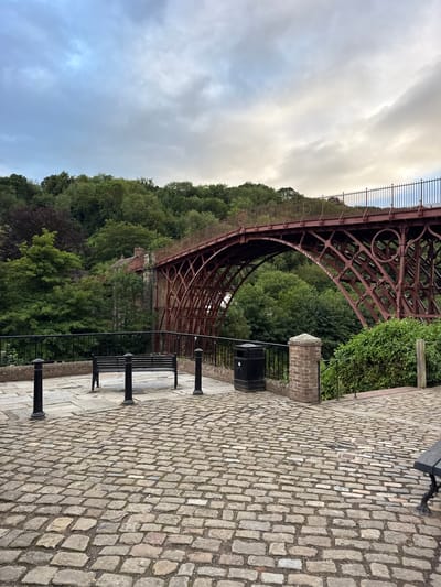 This is a photograph of the Iron Bridge in Ironbridge, United Kingdom, taken from a slightly elevated, paved viewpoint. The bridge, an iconic cast iron structure, spans a gorge and is painted a deep red. Its intricate lattice design is visible against a backdrop of lush green trees that line the gorge. The sky above is overcast with a mix of grey and white clouds. In the foreground, a cobblestone path leads towards the bridge. A black metal bench with a wooden seat is placed on the paved area, accompanied by several black bollards. A black trash bin is also situated on the paved area near a brick pillar. The overall scene depicts a historical landmark with a tranquil, natural setting.