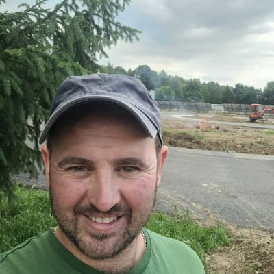 In Kaniów, Poland, a man wearing a dark blue baseball cap and a green t-shirt stands in front of a construction site. He is smiling, with a light beard and light brown eyes. Behind him, a small orange excavator is visible, along with a pile of dirt and some smoldering material. Lush green grass and trees frame the scene, with a cloudy sky overhead.
