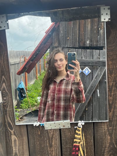 A woman takes a mirror selfie outdoors. She has long, dark, wavy hair and is wearing a red and brown plaid shirt over a tan t-shirt. She holds a smartphone in her right hand to capture the reflection. The background behind her is a rustic wooden fence and some greenery. The mirror itself is framed by weathered wood. The setting appears to be a yard or backyard, likely in Berezovskii, Russia, given the wooden structures and natural surroundings.
