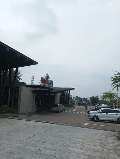 The image depicts the outdoor entrance area of an attraction identified by prominent signage as "MINI MUNDI TAMAN MINIATUR DUNIA," located in Indonesia, as indicated by the text and a visible red and white Indonesian flag. The scene is observed under an overcast, light grey sky.
The central feature is a modern-style building with a dark, angular, multi-faceted roof supported by dark, splayed beams. The building's facade on the left side is clad in light-toned stone, while the main entrance is a recessed, darker opening. Above this entrance, a large sign displays "MINI MUNDI" in stylized red and blue lettering with an accompanying graphic, and "TAMAN MINIATUR DUNIA" in smaller white text below. Behind the left portion of the building, a structure resembling cellular communication antennas is visible.
In the midground, a paved area functions as both an access road and a parking lot. A silver minivan is positioned beneath the covered entrance area. To the right of the entrance, several vehicles are parked; a silver SUV is most prominent in the foreground of this parking section. White arrow markings and a pedestrian crosswalk are painted on the asphalt surface.
Two individuals are visible: one person in dark attire is walking from right to left across the marked pedestrian crossing, and another individual wearing a light-colored shirt is partially visible near the left side of the building's entrance.
The immediate foreground consists of textured concrete pavement with a black drainage grate running horizontally. The background features numerous palm trees and other green foliage, with distant tree-covered slopes discernible beneath the horizon. A flagpole displaying the red and white flag of Indonesia is situated on the far right.