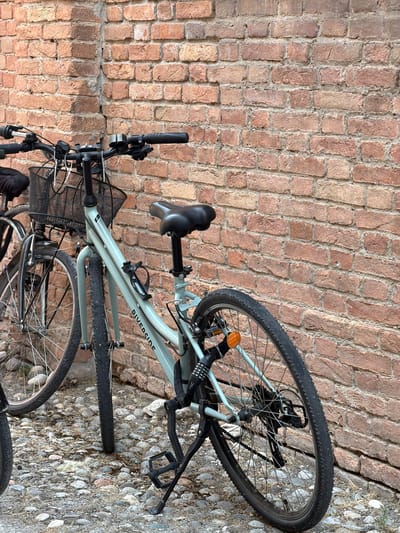 Two bicycles are parked against a brick wall in Piacenza, Italy. The bicycle in the foreground is a light blue hybrid bike with a black basket on the handlebars, a black seat, and a black U-lock secured to the rear frame. The brand "RIVERSIDE" is visible on the frame. The bicycle's kickstand is extended. The ground is covered with cobblestones. The brick wall behind the bicycles is made of red bricks with visible mortar. The overall lighting suggests it is daytime. There are no people visible in the image.