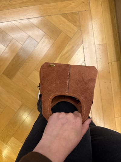 A high-angle shot shows a person sitting, holding a brown suede bag with a round handle. The person wears dark gray or black jeans. The background is a parquet wood floor, a light, natural tone. No details suggest Sunrise, United States of America.