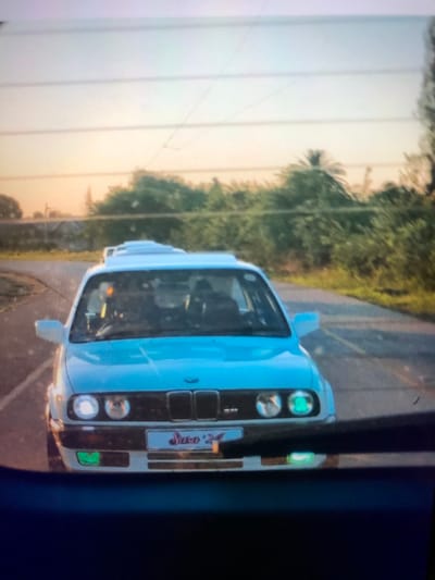 An undated, forward-facing photograph captures a white BMW E30 sedan on a paved road in what appears to be a semi-urban or peri-urban area within Soweto, South Africa, during either sunrise or sunset. The car occupies the central foreground, angled slightly, with its front fascia directly facing the viewer. Its left inner headlight is illuminated white, while its right inner headlight displays a distinct green illumination. Green-tinted fog lights are visible below the main headlight clusters. A license plate, partially obscured but appearing to read "Saso '27", is affixed to the front bumper. Two indistinct figures are visible through the windshield, one in the driver's seat and one in the front passenger seat. The car features a series of white, raised rectangular elements on its roof, consistent with multiple sunroofs or vents.
The vehicle is positioned on an asphalt road with visible lane markings, suggesting a slight curve. The background features dense green foliage and trees lining both sides of the road. A tall palm tree is discernible on the right side of the frame. In the distant left background, faint outlines of industrial structures, possibly silos or large buildings, are visible on the horizon. Multiple overhead power lines stretch horizontally across the upper portion of the sky, which transitions from a light orange-pink near the horizon to a pale blue overhead. The image appears to be taken from inside another vehicle, with a dark blue obstruction, possibly a dashboard or window frame, visible across the bottom fifth of the frame.