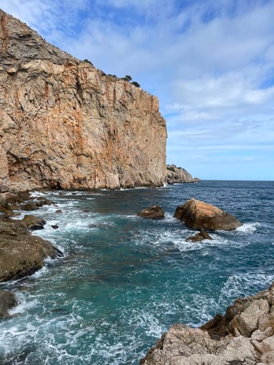 This image depicts a rugged coastal landscape in Torroella de Montgrí, Spain. A massive, vertical rock cliff, characterized by weathered tan, orange, and grey geological strata, occupies the left and central-left portions of the frame, extending from the bottom left upwards and receding into the distance. Sparse green vegetation is visible along the top edge of the cliff and on smaller landmasses further along the distant coastline. The Mediterranean Sea, exhibiting colors ranging from clear turquoise near the shore to a darker blue towards the horizon, fills the lower and right areas of the image. Waves are actively breaking against the base of the main cliff and numerous submerged and partially submerged rocky outcrops, generating white foam. Several prominent, reddish-brown and grey boulders are visible within the turbulent water. The sky above is partially cloudy, featuring a blue background with scattered white cumulus clouds. No individuals, human-made structures, or animals are present.