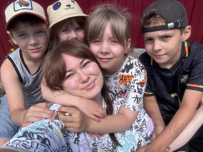 A group of five people, four children and one adult woman, are posing for a selfie outdoors. The woman is in the foreground, with her arms around two of the children. The children are of varying ages, and they are all smiling. One of the children is wearing a baseball cap with a bear graphic, another is wearing a straw hat with a panda design, and the third is wearing a black baseball cap. The adult woman has a tattoo on her arm. The background is a red wooden wall. The scene appears to be in a residential area, possibly a backyard.
