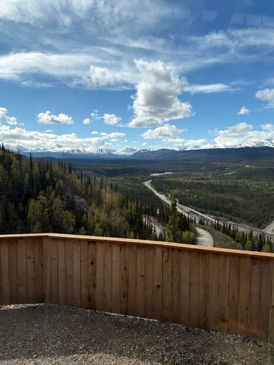 This landscape image, correctly oriented, captures a panoramic view of a vast natural wilderness, consistent with Denali National Park and Preserve. The shot is taken from an elevated viewpoint, with a sturdy wooden fence forming the lower edge of the frame, separating the scenic vista from a gravel-covered area in the immediate foreground.

Beyond the fence, a sprawling, dense coniferous forest dominates the midground, stretching across a wide valley. A paved road, characteristic of national park infrastructure, winds its way through the forested terrain, appearing to follow the path of a river or stream that is partially visible. On the right side of the road, nestled among the trees, a distinctive large, light-colored, dome-shaped structure, possibly a snow shed or a tunnel portal, stands out.

In the background, a majestic range of snow-capped mountains rises, their peaks sharp against the sky. These towering summits extend across the horizon, showcasing their rugged, snow-dusted surfaces. The sky above is a brilliant blue, adorned with a scattering of white clouds; some are fluffy and distinct, while others are thin and wispy, indicating clear and sunny weather.

The scene is illuminated by bright daylight, suggesting it is late morning or early afternoon. There are no visible people or discernible text in the image. The overall impression is one of grandeur, tranquility, and the awe-inspiring scale of nature.