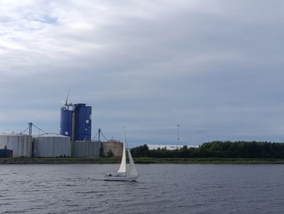 This media file captures a scene at Oulu, Finland, on a cloudy day. The image features a body of water in the foreground, with ripples suggesting a gentle breeze. On the water, a white sailboat with its sails up is being navigated by at least one person.
In the background, an industrial area is visible along the shoreline. Dominating the left side of the frame is a tall, blue cylindrical structure with white circular accents. Next to it are several large, light-colored storage tanks, some appearing metallic and others possibly concrete. Conveyor belts connect some of these structures. Further to the right, a long, low white building with a blue logo or text can be seen, partially obscured by a dense line of green trees that forms the natural backdrop to the industrial elements. A tall communication tower stands behind the trees to the right of the white building.
The overall mood is somewhat subdued, consistent with the overcast sky. The time of day is likely daytime, given the ambient light, but the cloud cover prevents strong shadows. The presence of the industrial facilities and the sailboat on the water suggests a working port or harbor environment within or near Oulu. No specific emotions are evoked, but the scene depicts a typical day of activity in a coastal industrial setting. Some text is visible on the tall blue structure, appearing in reverse and upside down as "TRBL OLCOTZ TRY".