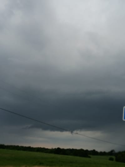 The image shows a landscape under a dark, cloudy sky. In the foreground, there is a field of green grass and a line of dark trees in the distance. A power line stretches horizontally across the scene, with a blue sign visible in the upper right corner. The sky is filled with heavy, gray clouds, suggesting an impending storm. The location is Waddy, United States.