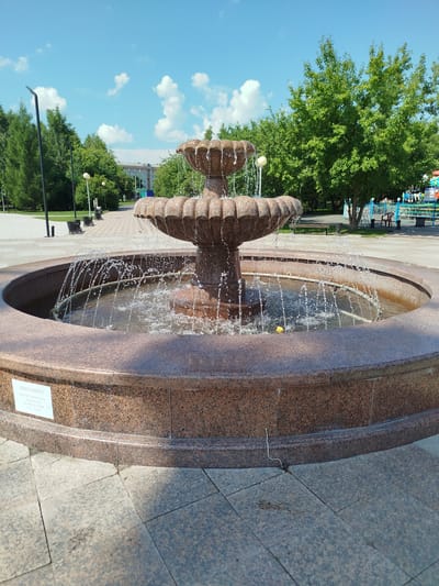 A multi-tiered granite fountain with water spraying from its basins is the central focus of this daytime outdoor scene in Yalutorovsk, Russia. The fountain is situated on a paved plaza, bordered by granite. Behind the fountain, a paved walkway leads past park benches and lampposts towards a distant building with light-colored walls and a darker roof. Lush green trees line the walkway and the background. A children's amusement area with colorful rides and fencing is visible to the right. The sky is a clear blue with scattered white cumulus clouds. A small white sign with text in Cyrillic is affixed to the front of the fountain's base.