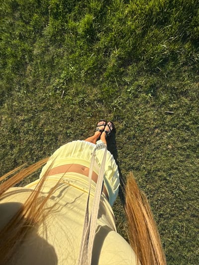 The image shows a person's lower body from a high angle, standing on grass. The person is wearing black sandals and a light yellow outfit, possibly a skirt and top, that exposes the midriff. Long, reddish-brown hair is visible, falling across the person's body. A beige strap from a bag crosses the front of the outfit. The background is a field of green grass, appearing somewhat uneven with scattered dry blades and twigs. The lighting suggests it is daytime with sunlight casting shadows, indicating an outdoor setting. The image does not contain information to identify the location as Bournemouth, United Kingdom.