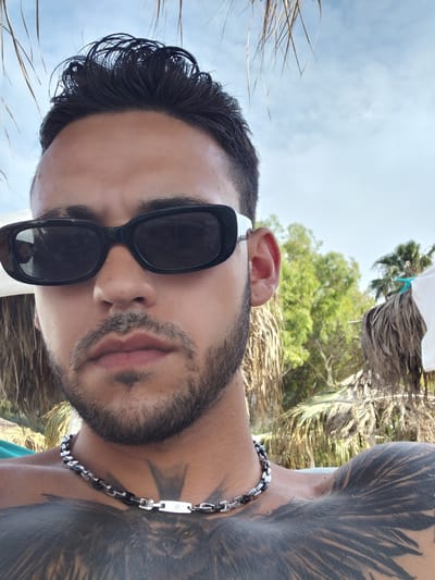 The image is a close-up, selfie-style shot of a man, oriented sideways. He is wearing black sunglasses and has a beard and mustache. He has dark hair and a large tattoo on his left shoulder and chest. He is also wearing a silver and black chain necklace. The background suggests an outdoor setting with green foliage and a bright sky, hinting at a sunny day. The man's expression is neutral, with his mouth slightly open.

The setting appears to be a beach or a similar tropical or Mediterranean location, possibly in Diakopto, Greece, given the mention of palm-like fronds and green vegetation against a clear blue sky. The lighting indicates it is daytime, likely sunny given the clear sky and the way light hits the skin and hair. There are no explicit signs of a specific event taking place; it appears to be a casual personal photograph. No visible text is present in the image.