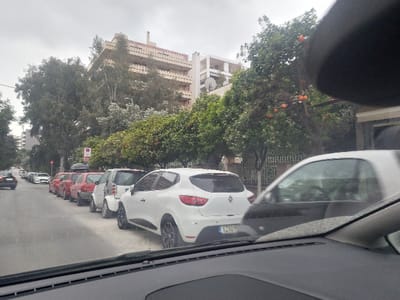The image presents an urban street scene in Nea Smirni, Greece, captured from within a vehicle looking forward through the windshield. The lower foreground shows the vehicle's dashboard and the base of the windshield.
On the right side of the street, a line of vehicles is parked. Closest to the camera is the rear of a white Renault Clio with a partial license plate "IZH 65xx" and multi-spoke black wheels. Behind it, a white and grey Smart Fortwo is parked. This is followed by a series of red hatchback cars, one of which features a black roof rack. Beyond the parked cars, a row of trees with orange fruit is visible, interspersed with taller, leafy trees. A decorative metal fence runs along the sidewalk behind the trees. A white pole with a circular sign (red ring, white background) stands above the parked cars.
The paved street extends into the distance, with a lane of moving traffic on the left, where several cars are visible, receding from the viewer.
In the background, a multi-story building with a distinctive stepped or terraced architectural design stands prominently, partially obscured by dense foliage. The sky is overcast. No individuals are visible in the scene.