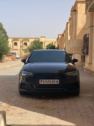 A black Audi A3 sedan is parked on a cobblestone street in front of a light tan building with arched windows and green tiled roofs. The car's front license plate reads "117693 119 16" with a German flag and the letter "D". The street is lined with trees and other buildings. The scene appears to be in the city of Boughezoul, Algeria, based on the architectural style. The sky is clear and bright, indicating daytime. No people are visible in the image.
