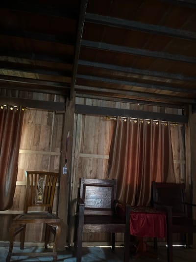 The image depicts the interior of a wooden room, likely a traditional house in Turumame, Indonesia. The walls and ceiling are constructed from wooden planks, with exposed wooden beams supporting the roof. On the left, a dark brown curtain with gold grommets hangs from a rod. A wooden chair with a slatted back and a carved detail in the center is positioned in front of the curtain. To the right of this chair, a second, darker wooden chair is visible, and further to the right, a third chair. Between the two right chairs, a small table covered with a red tablecloth is present. The room is dimly lit, with some light filtering through the left curtain, casting shadows on the wooden floor. A lanyard with a small tag hangs from a vertical wooden support in the center of the room.