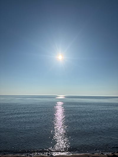 The image shows a serene seascape with the sun shining brightly in a clear blue sky. The sun's rays create a shimmering reflection on the calm water, which extends to the horizon. The foreground features a narrow strip of dark, pebbly beach. The scene evokes a sense of tranquility and natural beauty. The location of Turka, Russia, is not discernible from the visual information provided in the image.