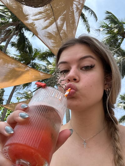 A close-up, eye-level selfie captures a young Caucasian female outdoors in an apparent resort or beach club setting in Thành phố Phan Thiết, Vietnam, under a bright, partly cloudy sky with visible palm trees and tan fabric shade structures overhead. The female subject, positioned centrally, has light brown hair, visible in a braid extending over her right shoulder (viewer's left). Her eyes are light-colored with prominent eyelashes. She is wearing small silver hoop earrings in her left ear (viewer's right) and a silver chain necklace with a small silver cross pendant. Her fingernails are painted a metallic light blue/grey. She holds a ribbed clear glass containing a reddish-orange beverage with ice, garnished with a sprig of dried greenery secured by a small orange clip. Her lips are pursed around an orange and white striped straw, creating a 'duck face' expression as she appears to be drinking. The background features a blue sky, dense green palm fronds, and the undersides of light-colored fabric awnings that exhibit shadows of palm leaves.