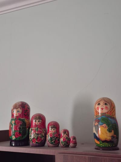 This media file captures a collection of matryoshka dolls displayed on a wooden shelf against a light-colored wall. The dolls are arranged in a line, decreasing in size from top to bottom. The largest doll, at the top, features a detailed painting of a woman with a colorful headdress and a scene with other figures. The subsequent dolls are smaller and display various painted motifs, primarily floral designs, with faces of women.
The setting appears to be indoors, possibly a home or a shop, given the presence of a shelf and wall. The lighting suggests it could be daytime, with ambient light illuminating the scene. There is no visible text in the image. The overall impression is one of traditional Russian craftsmanship and cultural display.