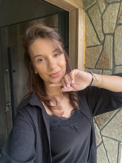 In a medium close-up, a young woman with medium-length brown hair and light skin poses for a selfie outdoors. She is wearing a black collared shirt over a black lace-trimmed camisole. Her left hand is raised, with her index finger touching her chin and her thumb tucked under her jaw. She is wearing a delicate gold heart pendant necklace and a bracelet on her left wrist. On her left forearm, a tattoo with Roman numerals is visible. She is standing in front of a door with a dark, textured surface and a stone wall. The setting appears to be the exterior of a building in Momchilovtsi, Bulgaria, evidenced by the architectural style of the door and the natural stone cladding. The lighting is natural, suggesting it is daytime.
