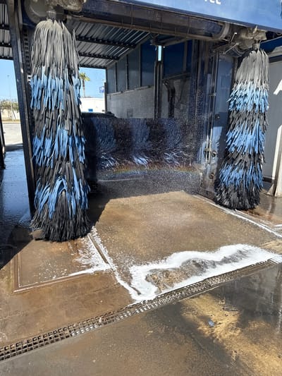 This image shows the interior of an automated car wash in Sollana, Spain. Two large, vertical car wash brushes with black and blue bristles are visible, one on the left and one on the right. Water is spraying from unseen nozzles, creating a misty effect and a rainbow on a dark surface behind the brushes. The ground in the car wash is made of a rough, textured material, with some areas covered in soap suds. A metal grate runs along the lower edge of the image. In the background, the roof of the car wash structure is visible, along with palm trees and a building under a clear blue sky.