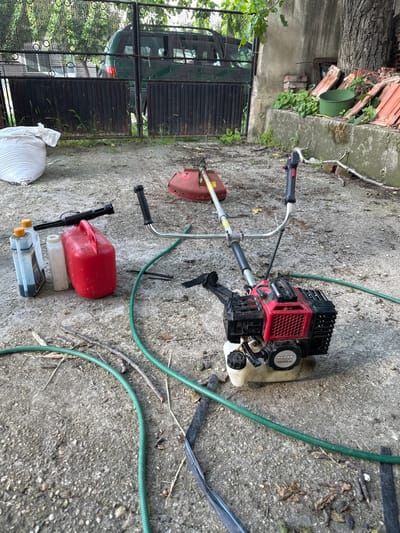 The image displays an outdoor concrete yard in Aidemir, Bulgaria, serving as a functional space for yard maintenance equipment. Centrally positioned is a disassembled red and black 'RAIDER RD-GBC20' gasoline brush cutter, with its engine unit and handlebars resting on the concrete, separated from its red cutting head which lies further back. Several lengths of green garden hose are coiled and spread across the concrete surface. To the left of the brush cutter's engine, a red plastic fuel can is present, alongside two clear plastic bottles containing a dark liquid and one translucent white bottle, accompanied by a black plastic nozzle or funnel. On the far left, a large white woven bag rests on the concrete. The concrete ground is dirty, speckled with small pebbles, patches of dirt, twigs, and dried leaves. In the background, a dark green metal fence, featuring circular perforations, partially obscures a dark green SUV and a backdrop of trees. To the right, a concrete wall forms a ledge supporting a pile of terracotta roof tiles, a green metal bucket, and some low-growing green vegetation. A substantial tree trunk is visible on the far right edge of the frame.