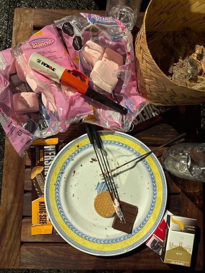 This image depicts a wooden slatted table in Huasca de Ocampo, Mexico, laden with items suggesting a recent s'mores preparation or consumption activity.
In the upper-left, an open, crumpled pink plastic bag of "Malvaviscos Bianchi" (marshmallows) contains several pink marshmallows. A white and orange Tokai long-stem lighter rests atop the bag. To its right, a woven straw basket holds wood kindling and debris. A clear glass is partially visible behind the marshmallow bag.
Centered on the table is a white ceramic plate with a yellow and blue patterned rim. On the plate, a single round biscuit and a rectangular piece of milk chocolate are present. Three metal skewers with black handles rest across the plate, one exhibiting white, melted marshmallow residue near its tip. The plate itself has numerous crumbs and smears of melted marshmallow and chocolate.
To the left of the plate, a "Hershey's King Size Milk Chocolate with Almonds" bar in its orange and brown wrapper is partially visible, alongside another Hershey's chocolate bar wrapper. To the lower right of the plate, a pack of Marlboro Gold cigarettes and a small yellow box labeled "DISFUNCIÓN ERÉCTIL" (Erectile Dysfunction) are visible. A clear plastic cup or glass is situated to the right of the plate. The overall scene appears untidy, characteristic of a casual, post-leisure activity. No individuals are present in the image.