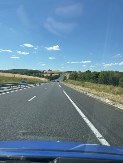 The image shows a view from inside a vehicle driving on a highway in a rural area. The highway stretches into the distance, curving to the right. The road is paved and has white lane markings. On the left side of the road, there is a metal guardrail and a grassy embankment with some trees and fields beyond. The fields appear to be harvested, with some dry grass or stubble visible. On the right side of the road, there is a grassy shoulder and more trees. The sky is blue with scattered white clouds. In the distance, there are rolling hills covered with trees and fields. Several vehicles are visible on the highway, appearing as small shapes in the distance. The image is taken from a low angle, suggesting it was captured from the dashboard or windshield of the car. The blue hood of the vehicle is visible in the foreground, slightly out of focus. The scene is consistent with a highway drive through the French countryside, specifically in the area of Villers-Agron-Aiguizy, France, known for its rural landscapes and agricultural fields.