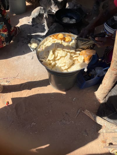 The image depicts an outdoor food preparation and vending scene in Vodni, Nigeria. In the foreground, a large, galvanized metal bucket is filled with numerous light-colored, oval-shaped fried snacks. On top of these, a smaller quantity of darker, golden-orange fried snacks is visible. A person on the right, wearing a patterned garment, uses both hands to open a black plastic bag, with other packaged food items in clear plastic bags visible nearby, including one with an orange-yellow substance. To the right of the bucket, a blue plastic basket containing various small items in plastic wrapping sits on the reddish-brown earthen ground.

In the mid-ground, to the left of the main bucket, a smaller silver bowl contains additional light-colored fried snacks. Behind these, a rudimentary cooking setup is present, consisting of a black pot elevated on several stones, with light-colored items frying within the pot over what appears to be an open fire or embers.

On the far left, the lower legs and feet of a person wearing a dark, patterned fabric are visible on a lighter-colored ground surface, possibly concrete or packed earth. Strong sunlight casts distinct, elongated shadows across the reddish-brown ground, which contains scattered debris such as small sticks and wood fragments. A thick tree trunk or wooden post is visible on the far right side of the frame.