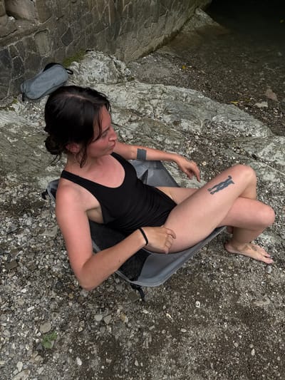 A woman with dark hair is lying on her back on a rocky surface near a body of dark water. She is wearing a black tank top and black shorts. Her legs are crossed, and she is lying inside a grey backpack. She has a tattoo of a diver on her right thigh. There is a small grey bag next to her head. The scene appears to be outdoors, possibly in a natural setting with rocks and water. The lighting suggests it could be daytime. There is no visible text in the image.