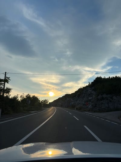 A POV shot from a vehicle driving on a winding road in Gornja Brova, Montenegro, at sunset. The sun is low in the sky, casting a warm glow and reflecting off the wet road. The road has white lane markings and is bordered by a rocky hillside on the right and vegetation on the left. A power line stretches across the sky. There are no people visible in the image. The overall scene is serene and picturesque.