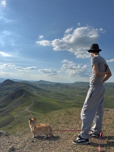 A man wearing a grey tracksuit and a black hat stands with his arms crossed, with a ginger and white corgi dog on a pink leash standing next to him. They are positioned at the edge of a grassy hill overlooking a mountainous landscape with a blue sky and white clouds above. The location is in or near the city of Орджоникидзевский поселковый совет, Ukraine, as the photo was taken there.