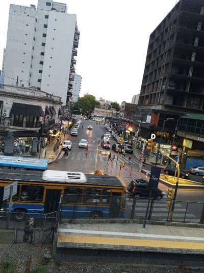 The image captures an elevated view of a bustling urban intersection in Ramos Mejía, Argentina, under an overcast late afternoon or early evening sky. In the immediate foreground, a metal fence and a concrete platform with yellow safety markings are visible. A blue and yellow articulated bus, displaying motion blur, occupies the lower center, moving from left to right. Two individuals are standing close together to the left of the bus, near the fence.
The street below is wet, indicating recent precipitation. Multiple vehicles, including cars and a dark SUV, are present; some are parked along the curbs, while others are in traffic. Pedestrians are observed on sidewalks and utilizing marked crosswalks. Traffic lights are operational, with one appearing red for cross-traffic.
To the left, a tall, white multi-story residential or office building with numerous dark-framed windows and balconies rises prominently. Below it are lower commercial buildings featuring dark awnings and illuminated storefronts. To the right, a dark, skeletal, multi-story concrete structure, appearing unfinished or derelict and covered in graffiti, dominates the right side of the frame. Adjacent to this structure are further commercial establishments, one displaying a large "D" sign and a "Luccas" banner. Overhead wires are visible across the scene.