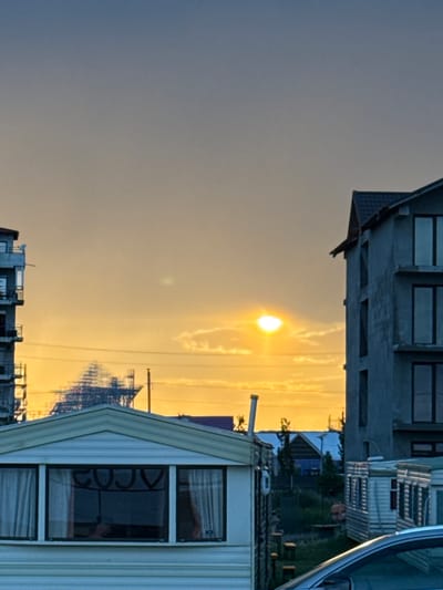 The image displays an urban or semi-urban scene in Costinești, Romania, during either sunrise or sunset, evidenced by the sky's gradient from deep blue-grey at the top to vivid orange and yellow near the horizon. The sun, a brilliant white-yellow disk, is visible in the upper mid-right, partially obscured by thin clouds.
In the mid-ground, a prominent, dark-toned, intricate scaffolding or metal structure is silhouetted against the illuminated sky. To the left, a multi-story building, appearing to be under construction, exhibits visible framework and concrete elements. On the right, a multi-story, unfinished concrete building with multiple dark, unglazed window openings occupies a significant portion of the frame. Below this structure, several white, rectangular prefabricated mobile homes are observed.
In the immediate foreground, a white, single-story mobile home with a distinctive gable roof is centrally positioned. Its front features three windows; the central window reflects inverted alphanumeric characters resembling "2020." Light-colored curtains are visible behind the windows. To the right, the silver roof and windshield of a car are partially visible in the bottom-right corner. Utility poles and horizontal power lines are present, traversing the mid-sky. Sparse vegetation, including grass and small trees, is visible between the various structures at ground level.