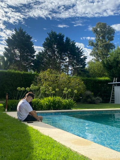 A person sits at the edge of a pool, facing away from the camera, dressed in a white shirt and dark shorts. The pool's edge is a light tan color and the pool itself is surrounded by lush green grass and various plants. Tall green trees tower in the background against a bright blue sky filled with fluffy white clouds.