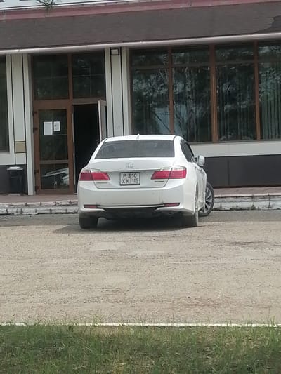 A white sedan is parked on a gravel lot in front of a building with large windows and a dark-tiled roof. The car is facing away from the camera, and its license plate is visible. The license plate reads "P 310 XK 125". The building appears to be a commercial or public building. The environment is outdoors, with some dry grass in the foreground. The location is the city of Lesozavodsk, Russia.